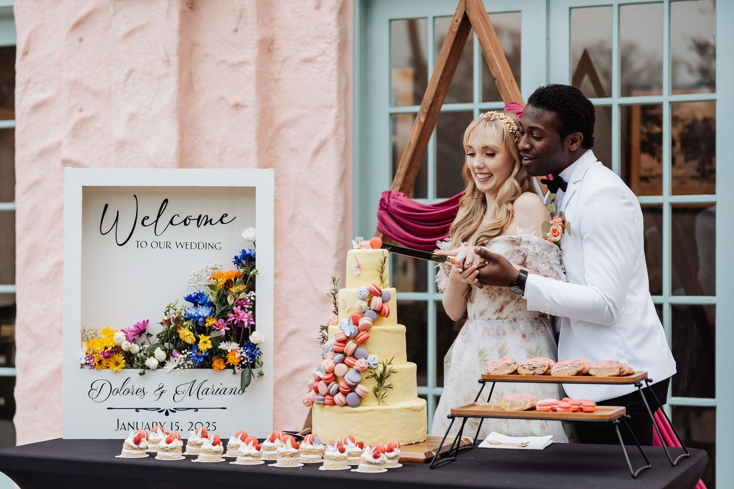 A couple shares a sweet moment cutting their wedding cake together at a reception dessert table with colorful floral decor.