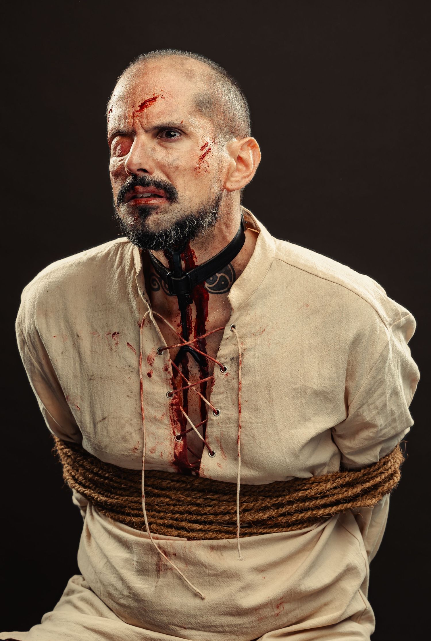 Dramatic theatrical photo of a prisoner in white clothing bound with rope showing signs of violence and distress.