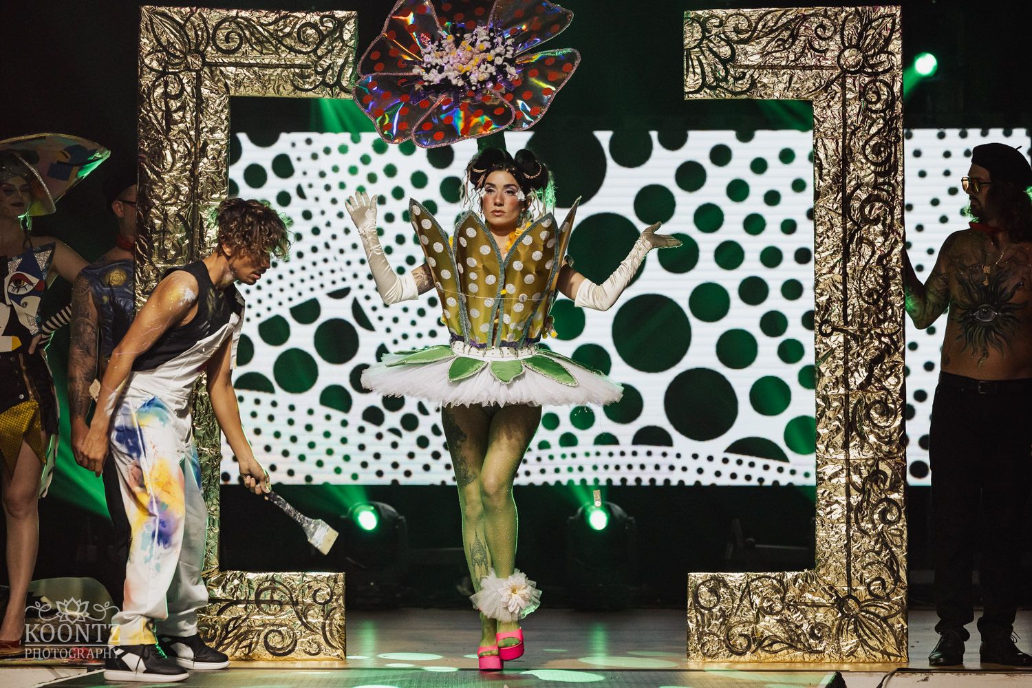 Fashion models display colorful outfits on a runway with polka dot backdrop and ornate gold-framed arches.