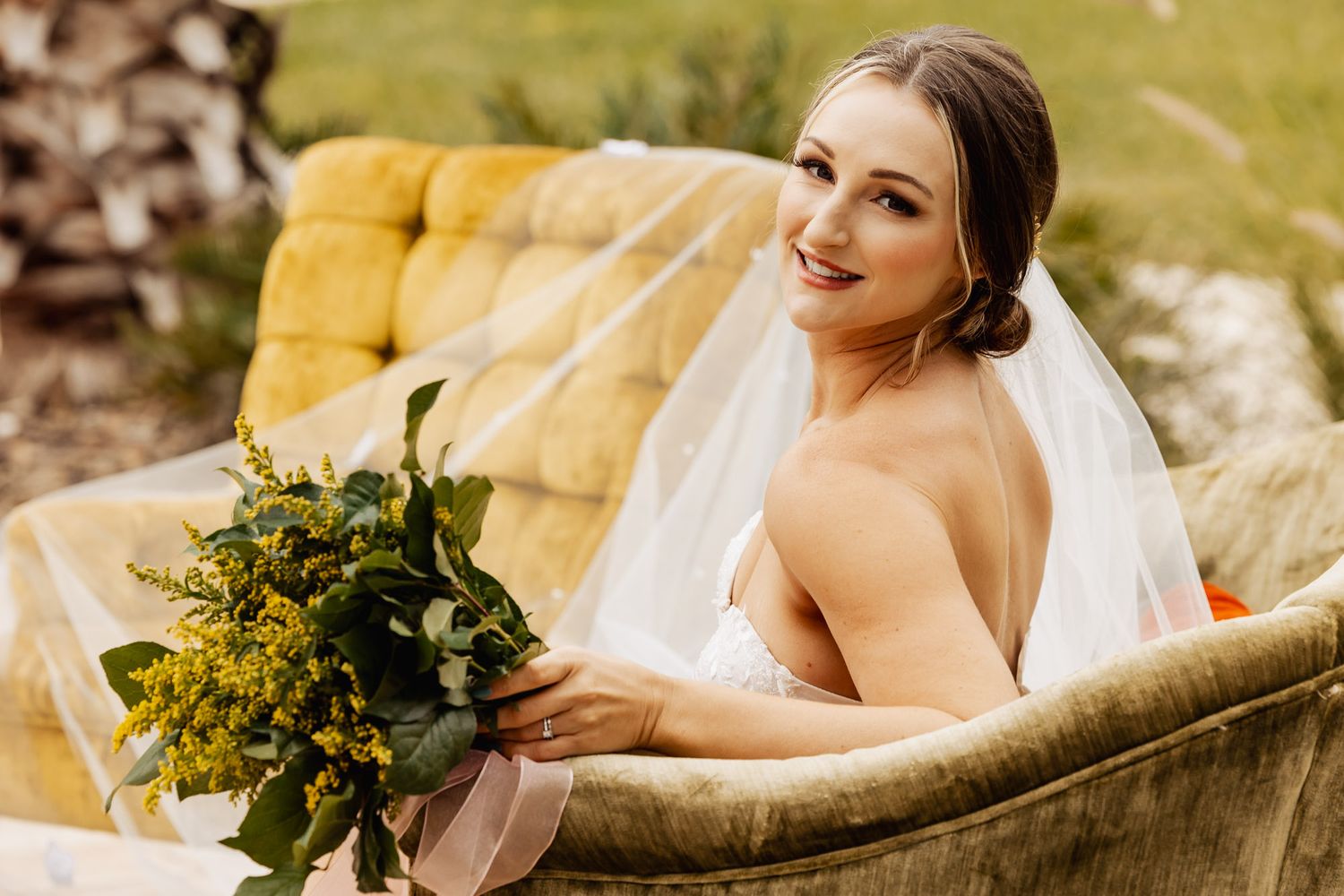 Smiling bride in white dress holds yellow and green bouquet while seated on a vintage yellow velvet sofa.