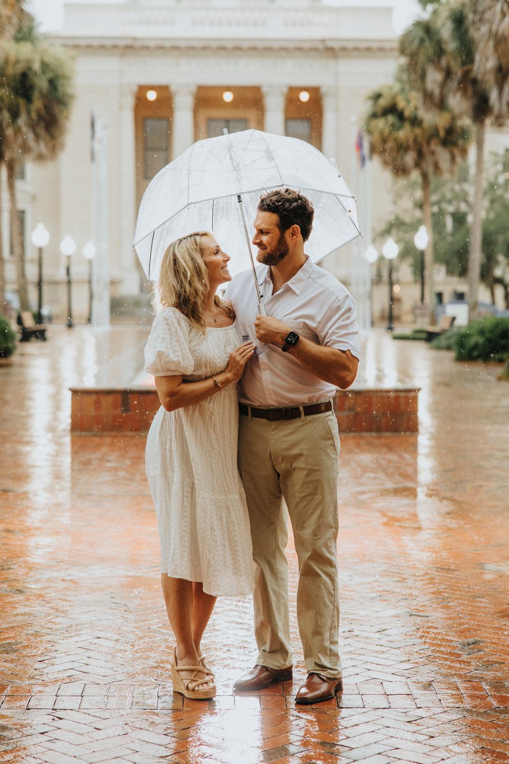 Romantic couple sharing an umbrella on rainy day near classical building with palm trees.
