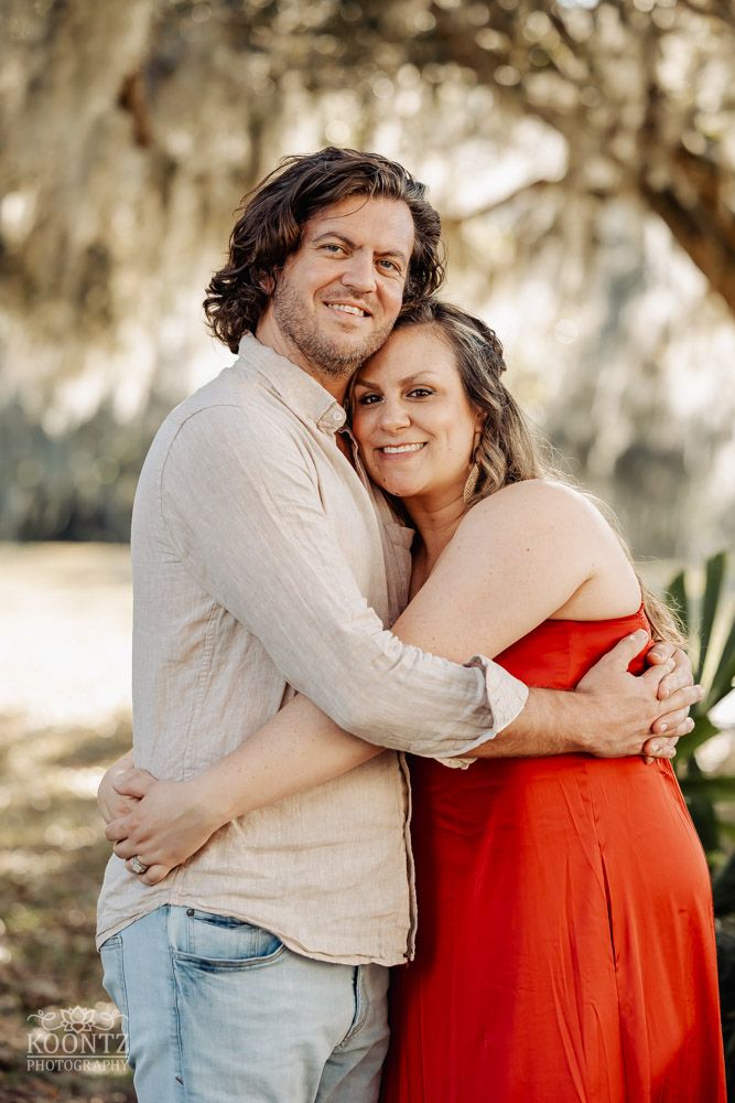 Outdoor portrait of couple embracing under mossy trees, woman in red dress and man in beige shirt.