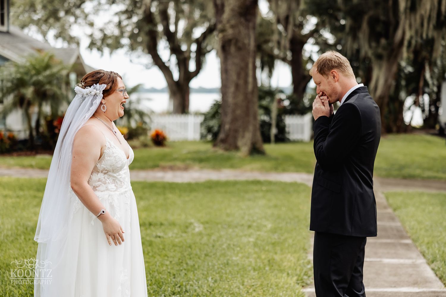 First look reaction between bride and groom on landscaped lawn with Spanish moss trees.