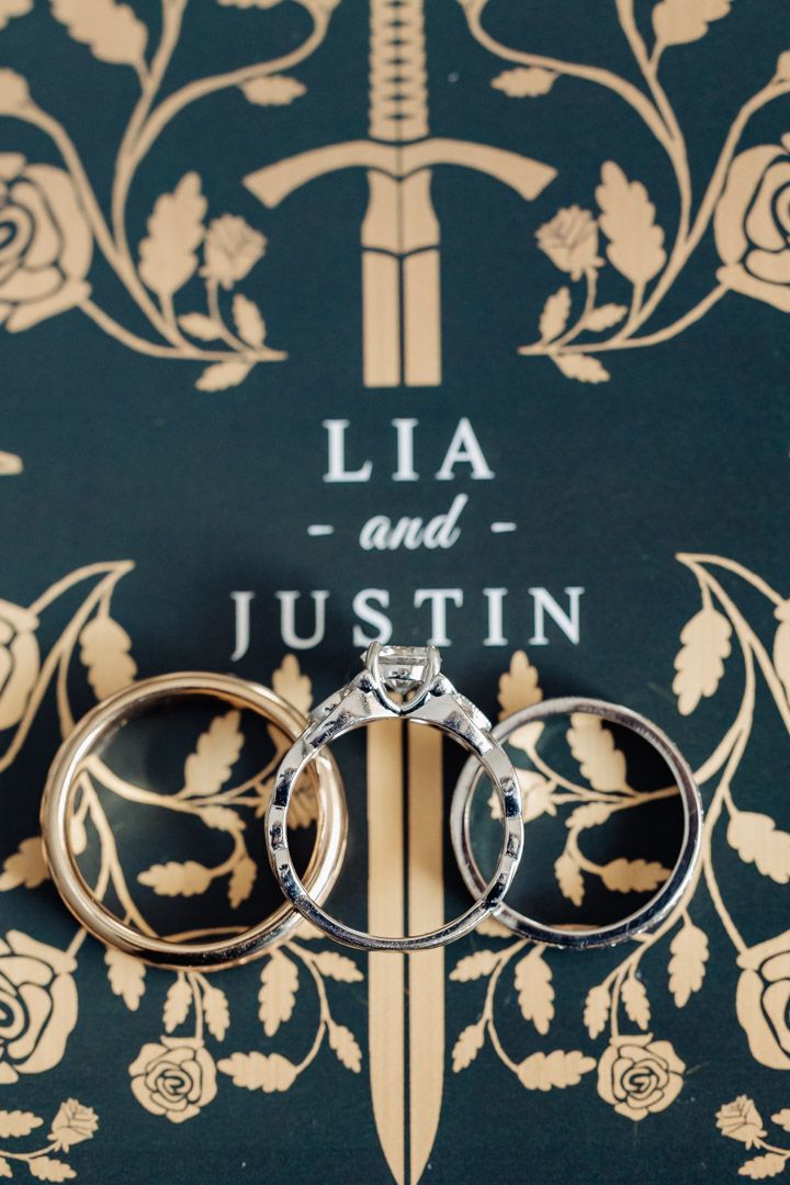 Three wedding rings displayed on a dark green book cover reading Lia and Justin with sword design.