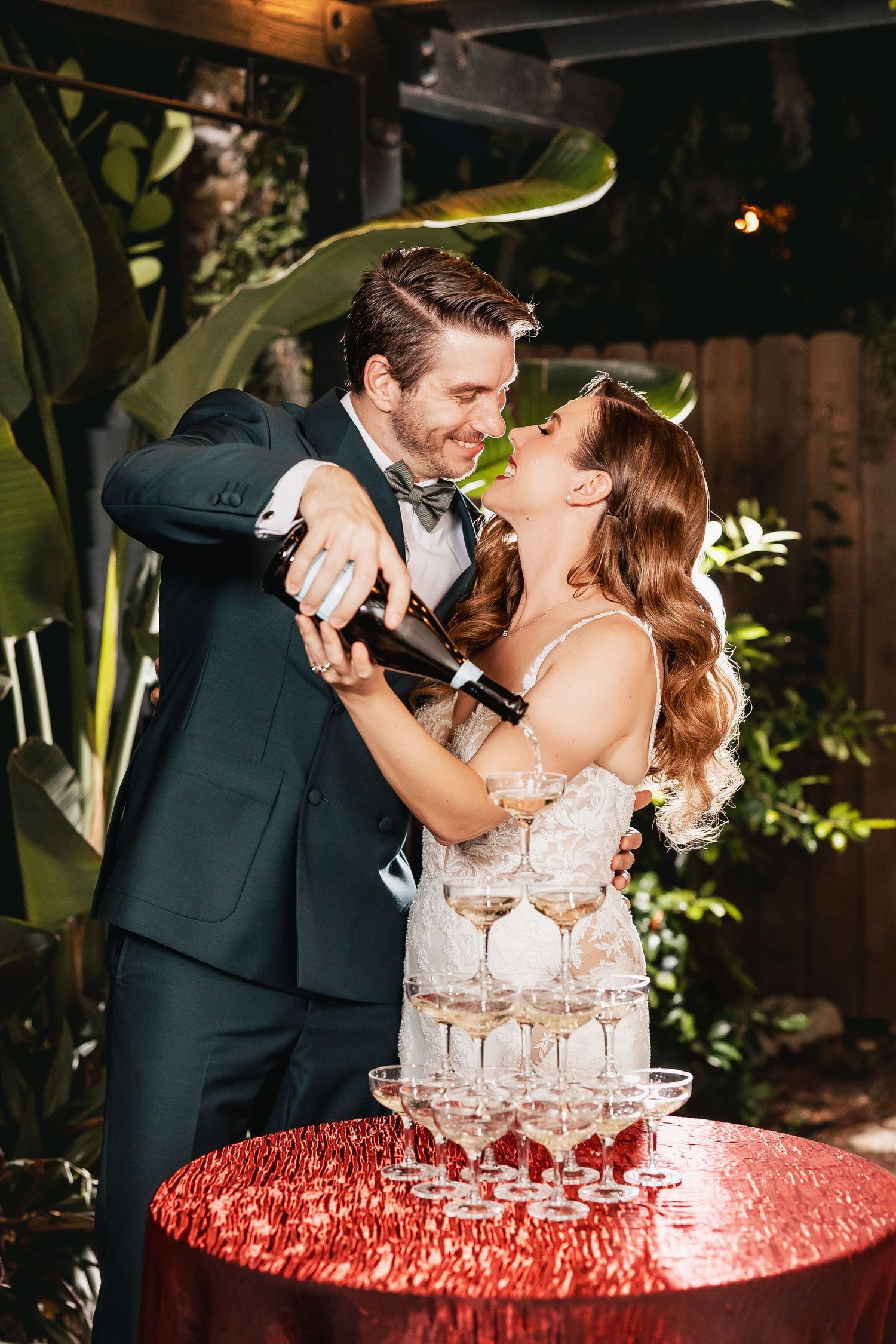 Bride and groom pouring champagne tower at wedding reception outdoors.