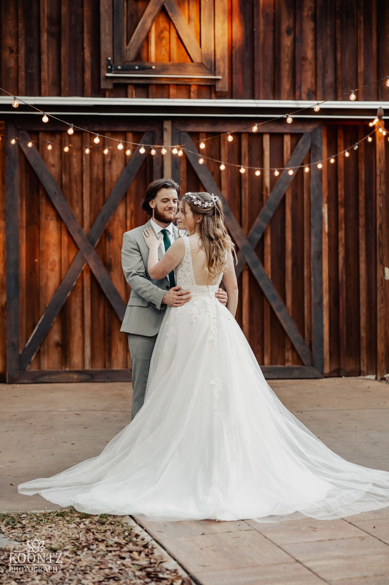 Bride and groom embrace romantically in front of rustic wooden barn adorned with string lights.