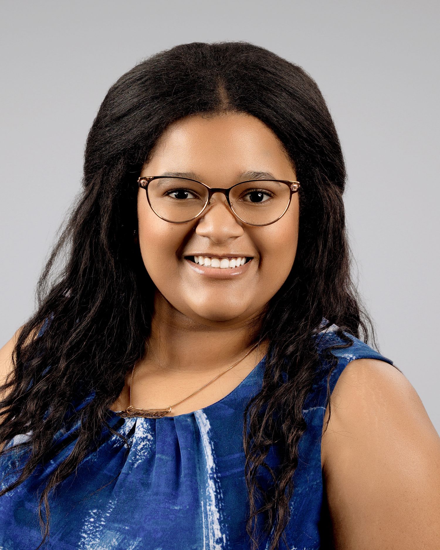 Young smiling woman with curly black hair and glasses wearing a blue sleeveless top.