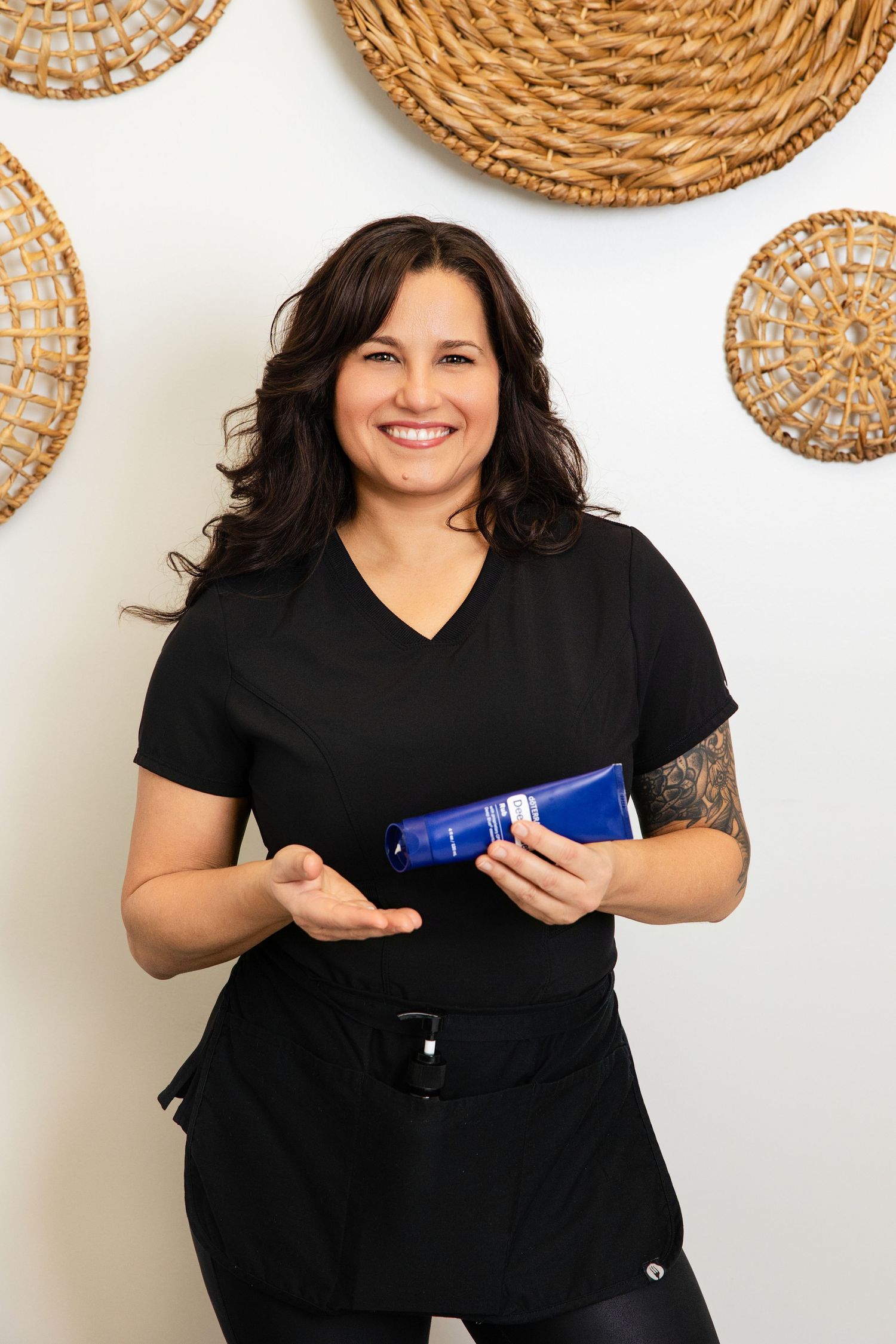 Smiling female esthetician in black scrubs holding a blue skincare product tube in a spa setting.