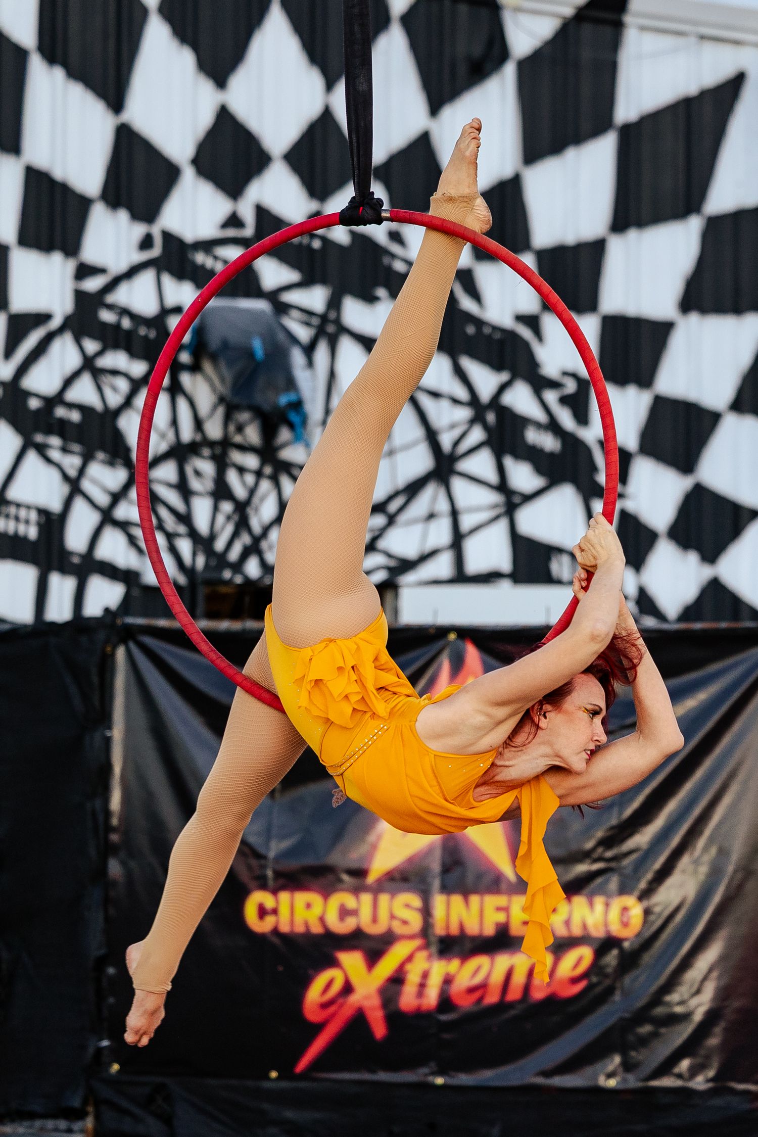 Flexible female aerialist in yellow shorts performing a split on a red aerial hoop at Circus Inferno.