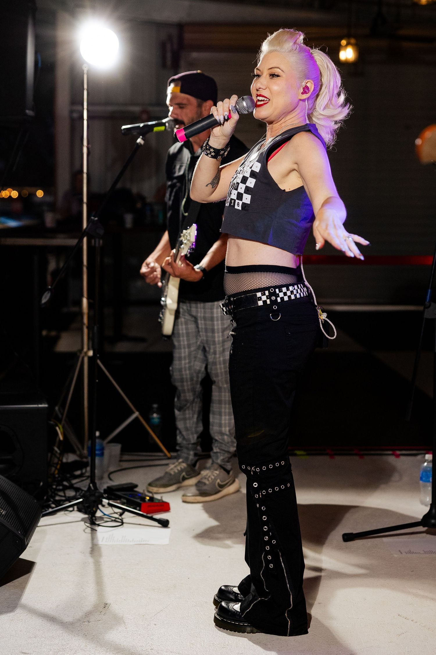 Female punk rock singer in black outfit and platform boots performing on stage with band.
