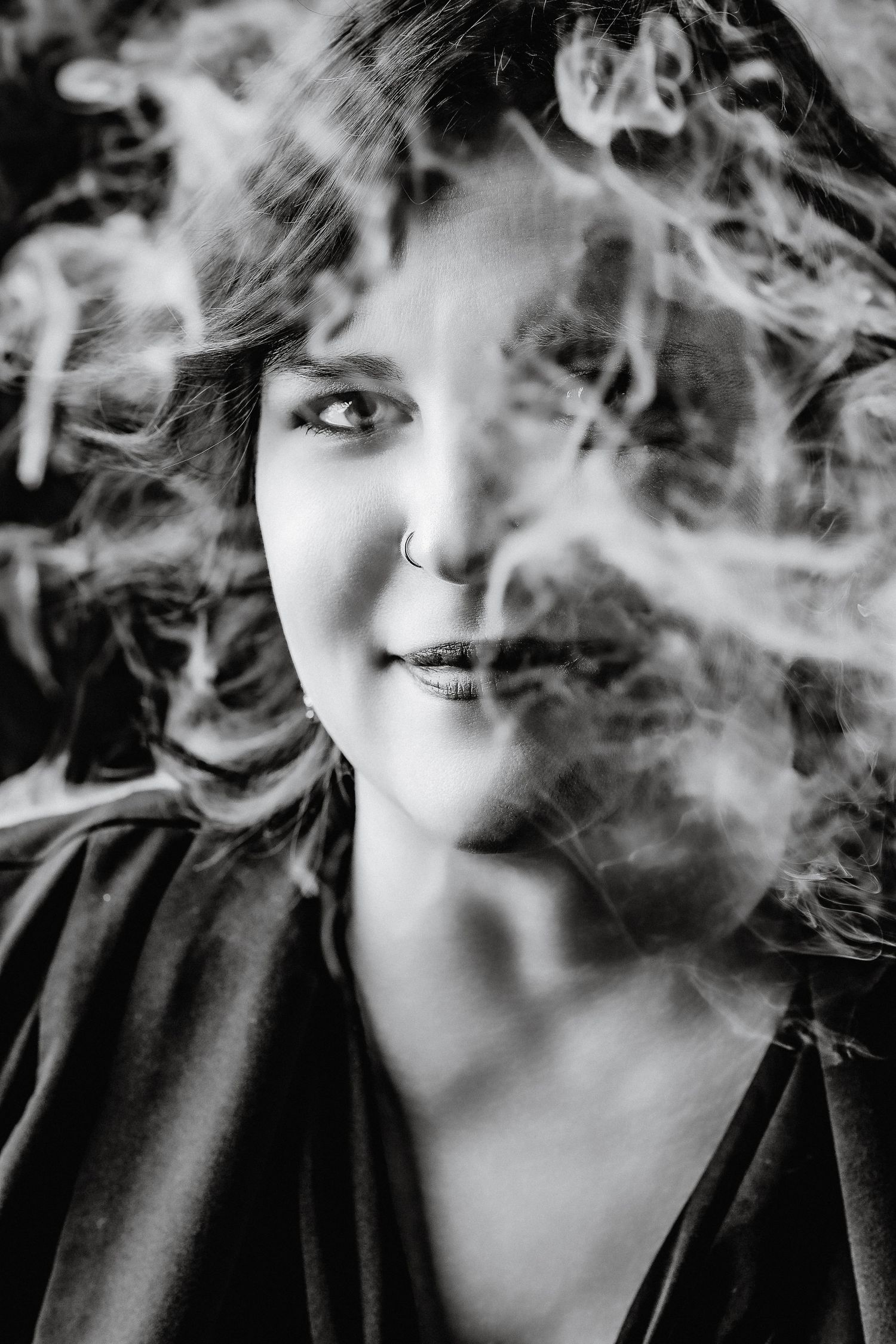 Black and white portrait of a curly-haired woman with nose ring surrounded by swirling smoke.
