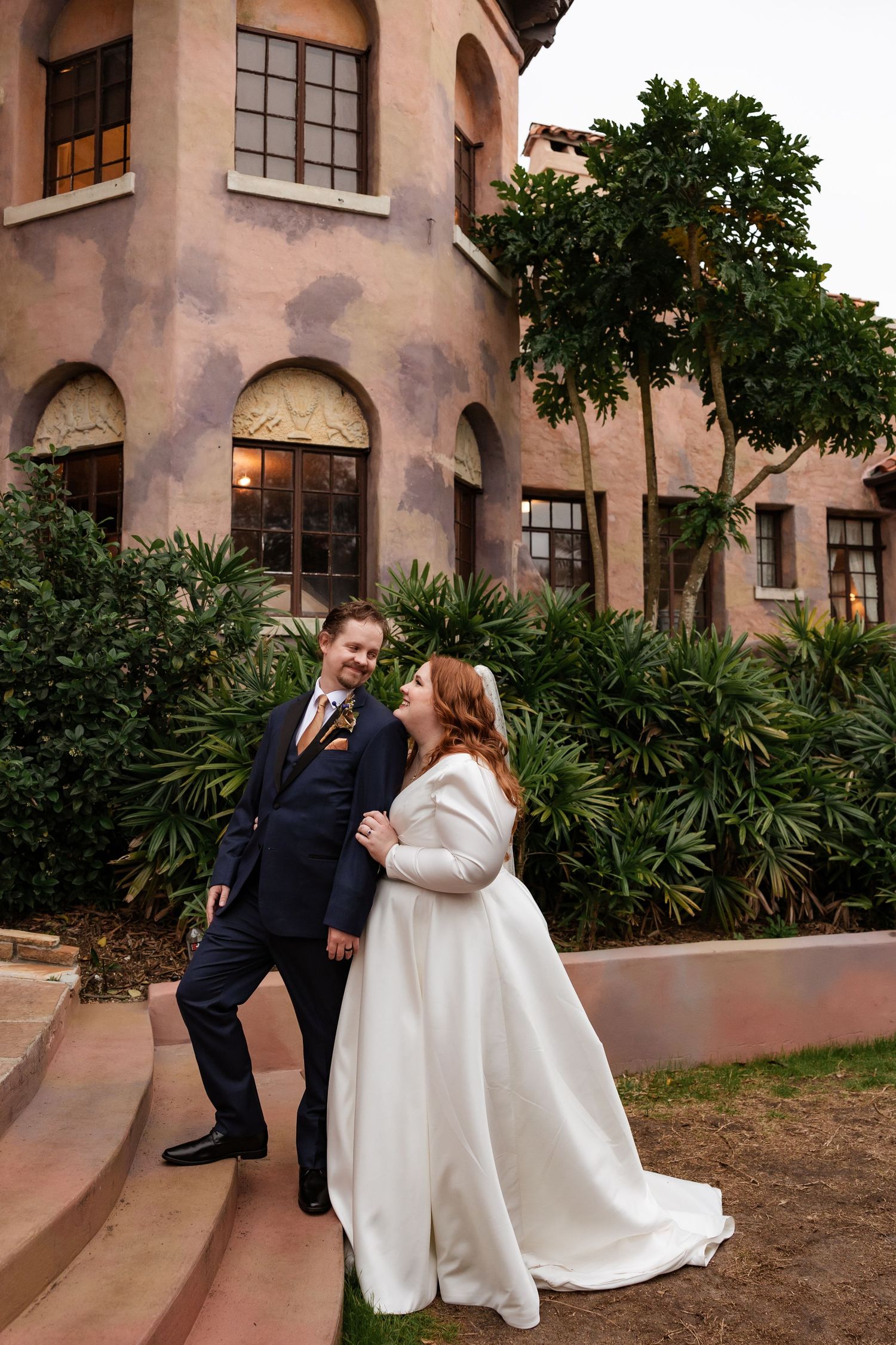 Bride and groom share a joyful moment on steps outside a historic Spanish-style building.