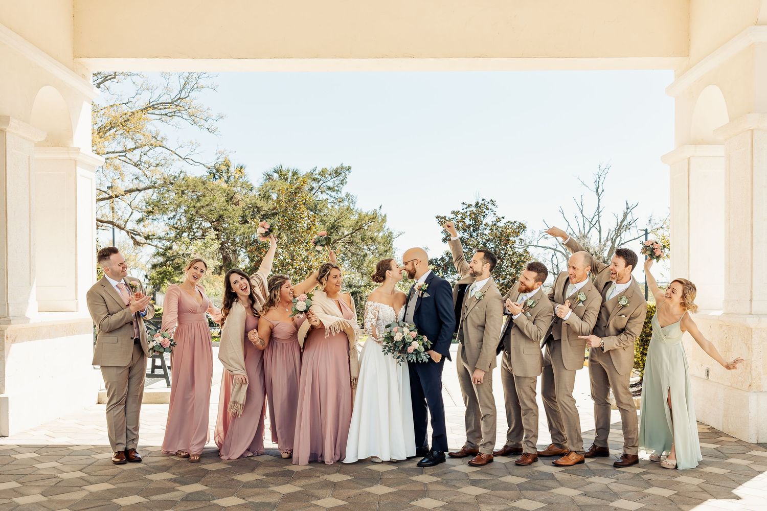 Wedding party celebrating under a portico with bridesmaids in mauve and groomsmen in tan suits.