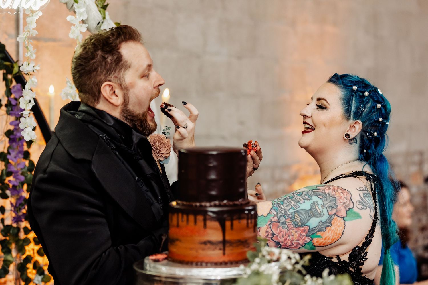 Laughing bride feeds groom a bite of dark chocolate wedding cake during fun cake cutting ceremony.