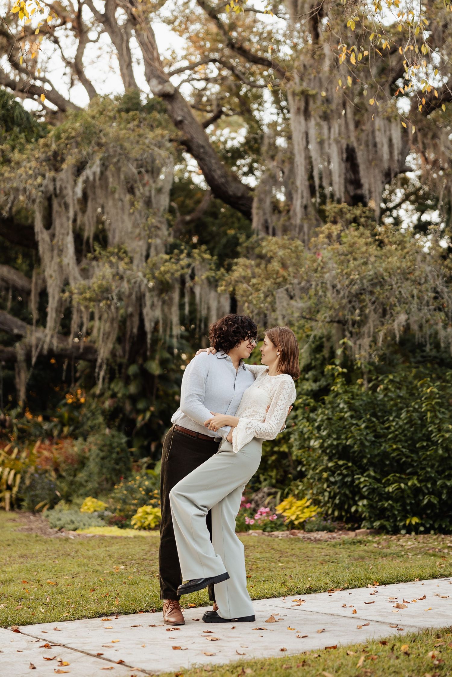 Two women dance and laugh together on a garden path beneath a large moss-draped oak tree.