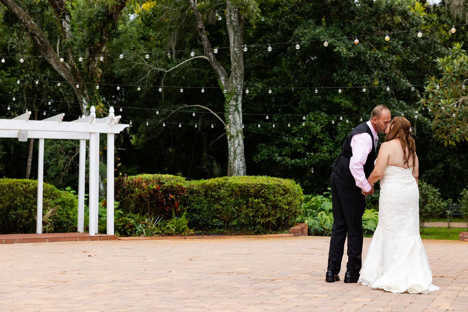 Bride and groom share a tender kiss on a brick patio with white arbor and string lights.