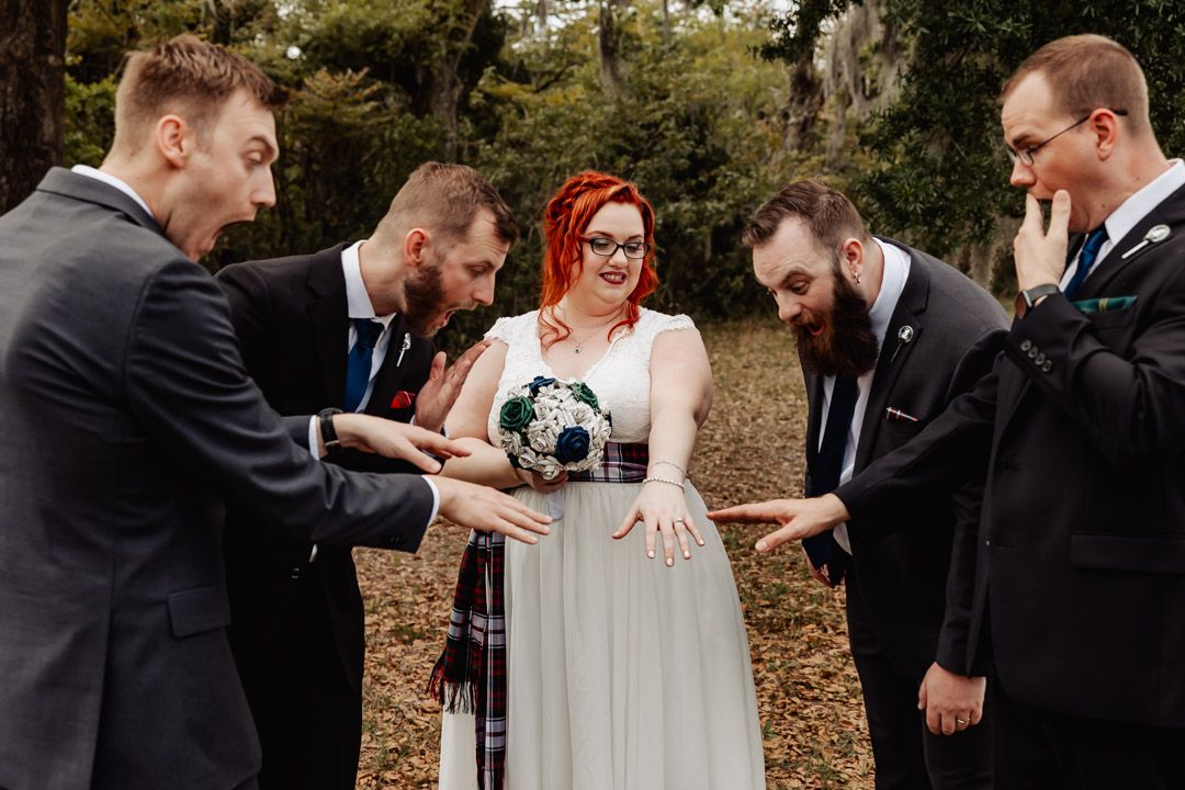 Bride and groomsmen gather in circle examining bouquet under moss-draped trees at outdoor wedding celebration.