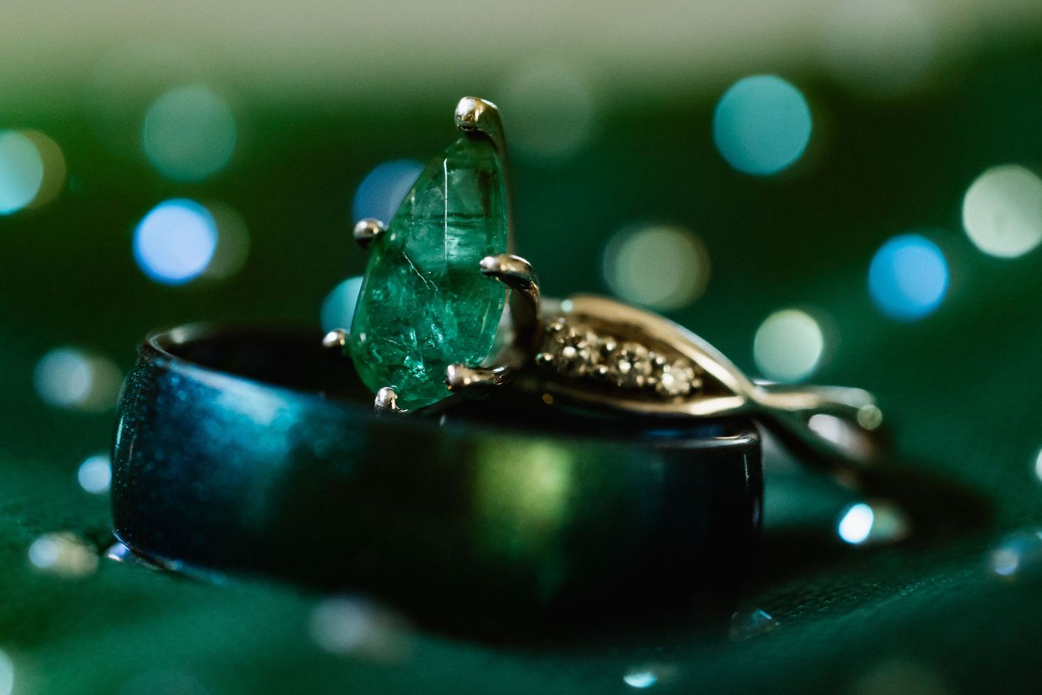 Close-up of an emerald gemstone engagement ring beside a dark wedding band on a green surface.