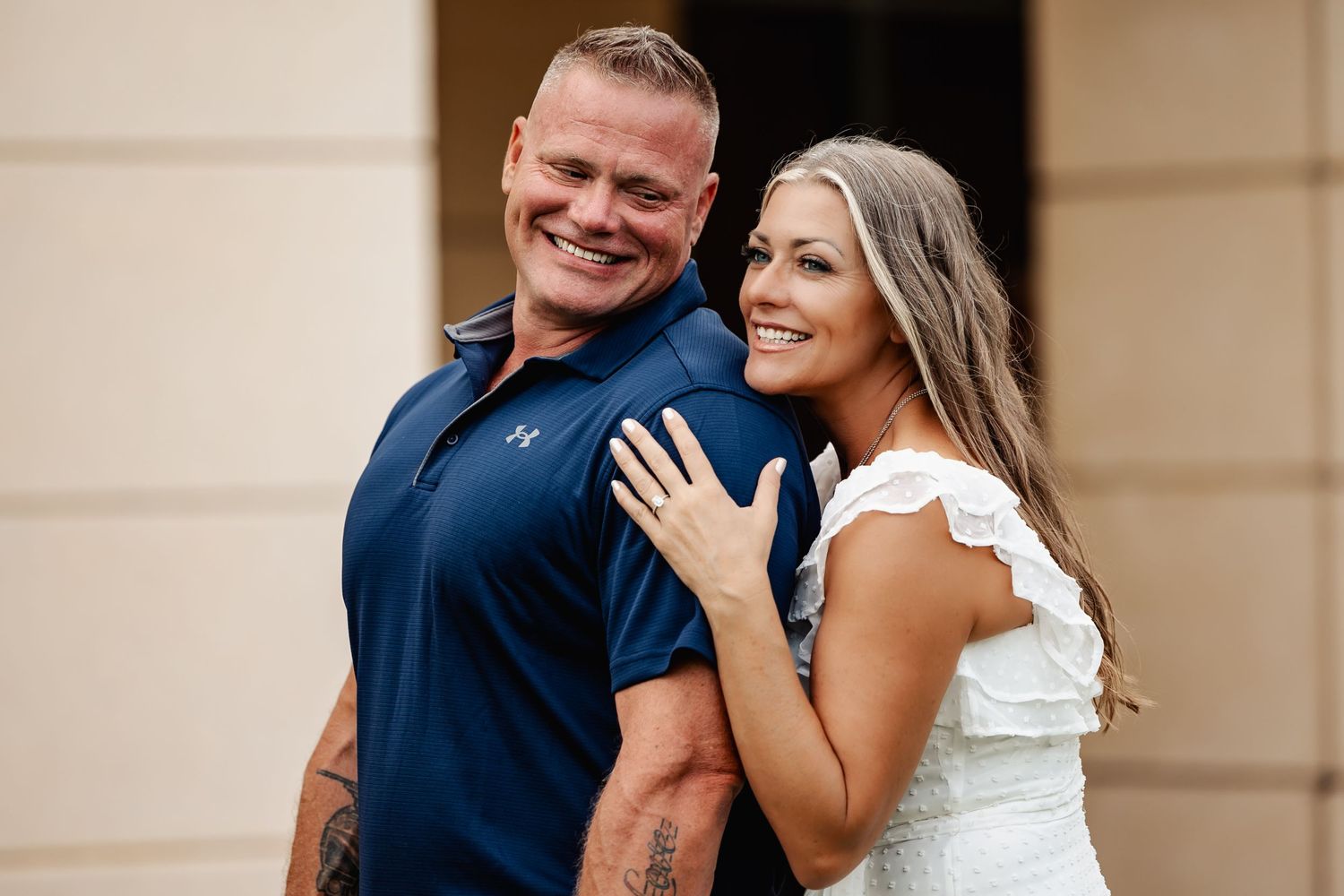 A joyful couple shares a candid moment while posing against a beige wall outdoors.