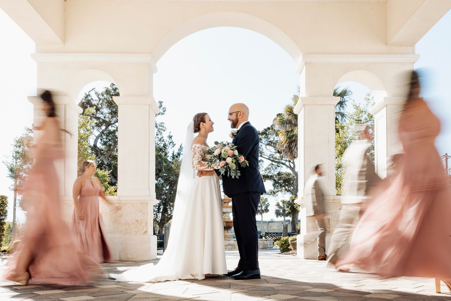 Elegant couple exchange vows under white archway as bridesmaids in blush dresses create motion blur effect walking by.