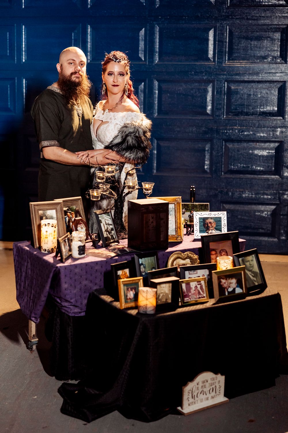 A couple poses behind a memorial display table decorated with vintage frames and purple velvet cloth.