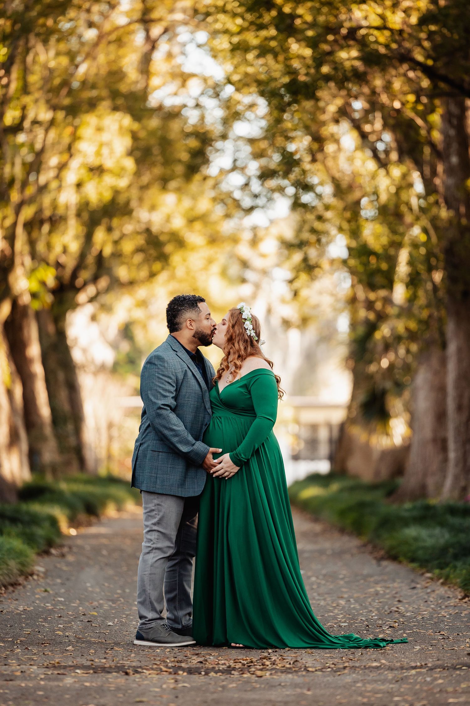 Couple sharing romantic moment on tree-lined path, pregnant woman in green maternity gown.