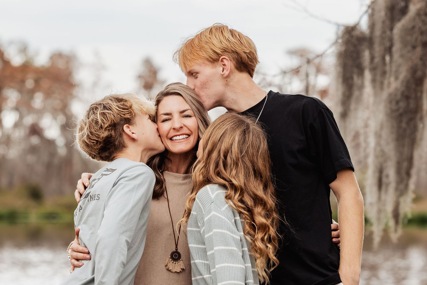 A happy mother laughs joyfully as her two teenage sons kiss her cheeks outdoors near a lake.