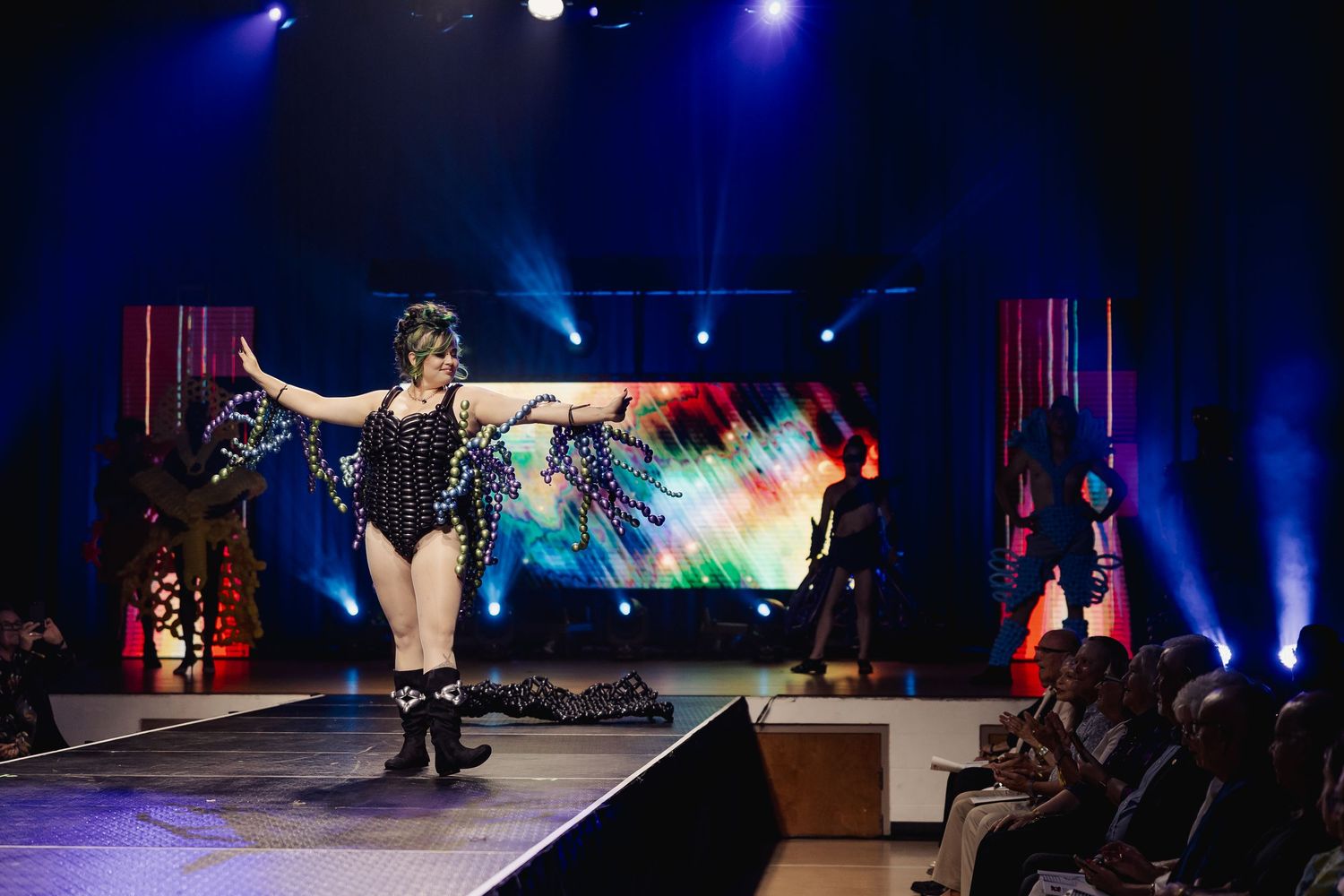 Model in black sequin outfit with balloon accessories performs on a lit runway before an audience.