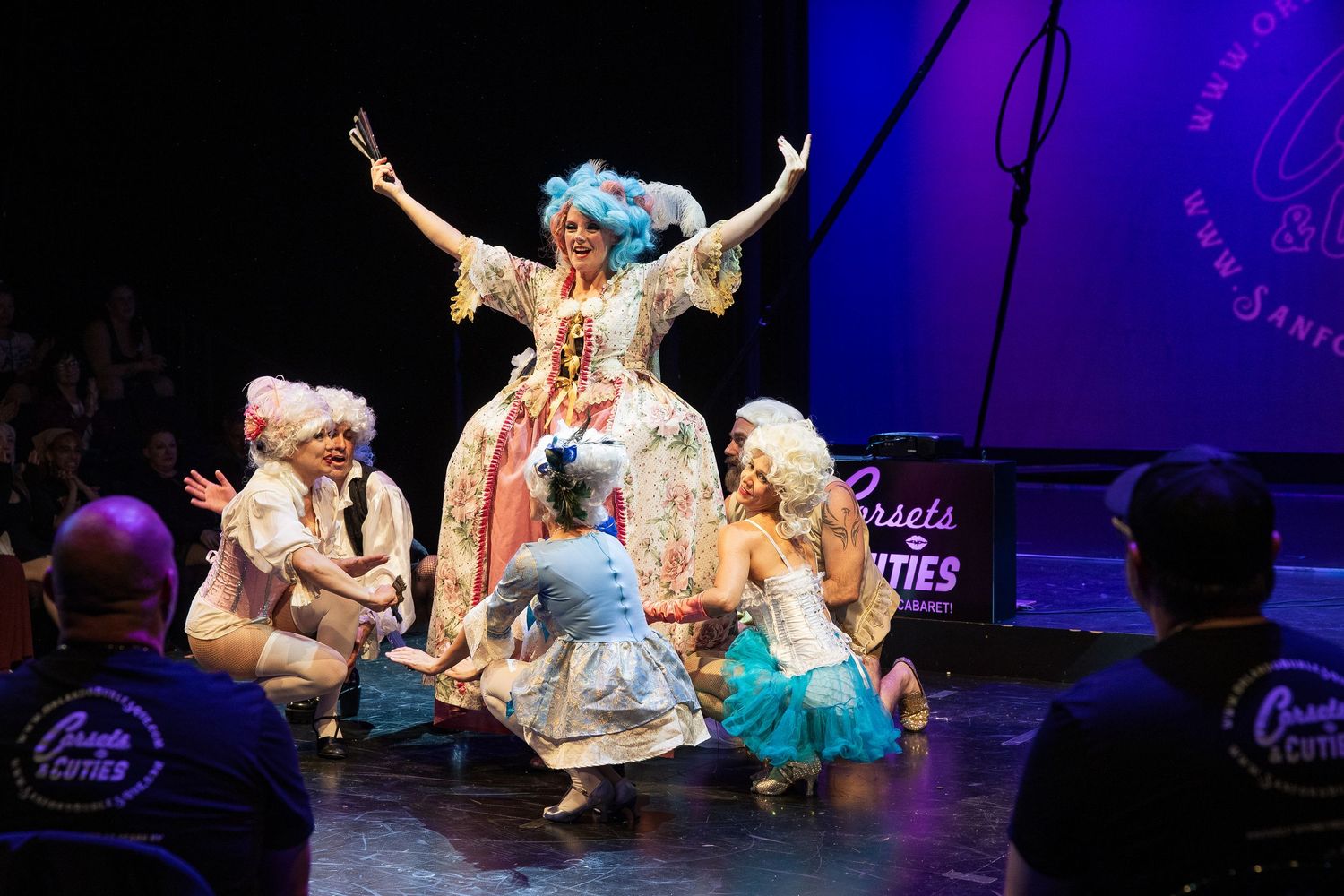 Burlesque performer in elaborate floral gown raises arms triumphantly while cast kneels around her on stage.