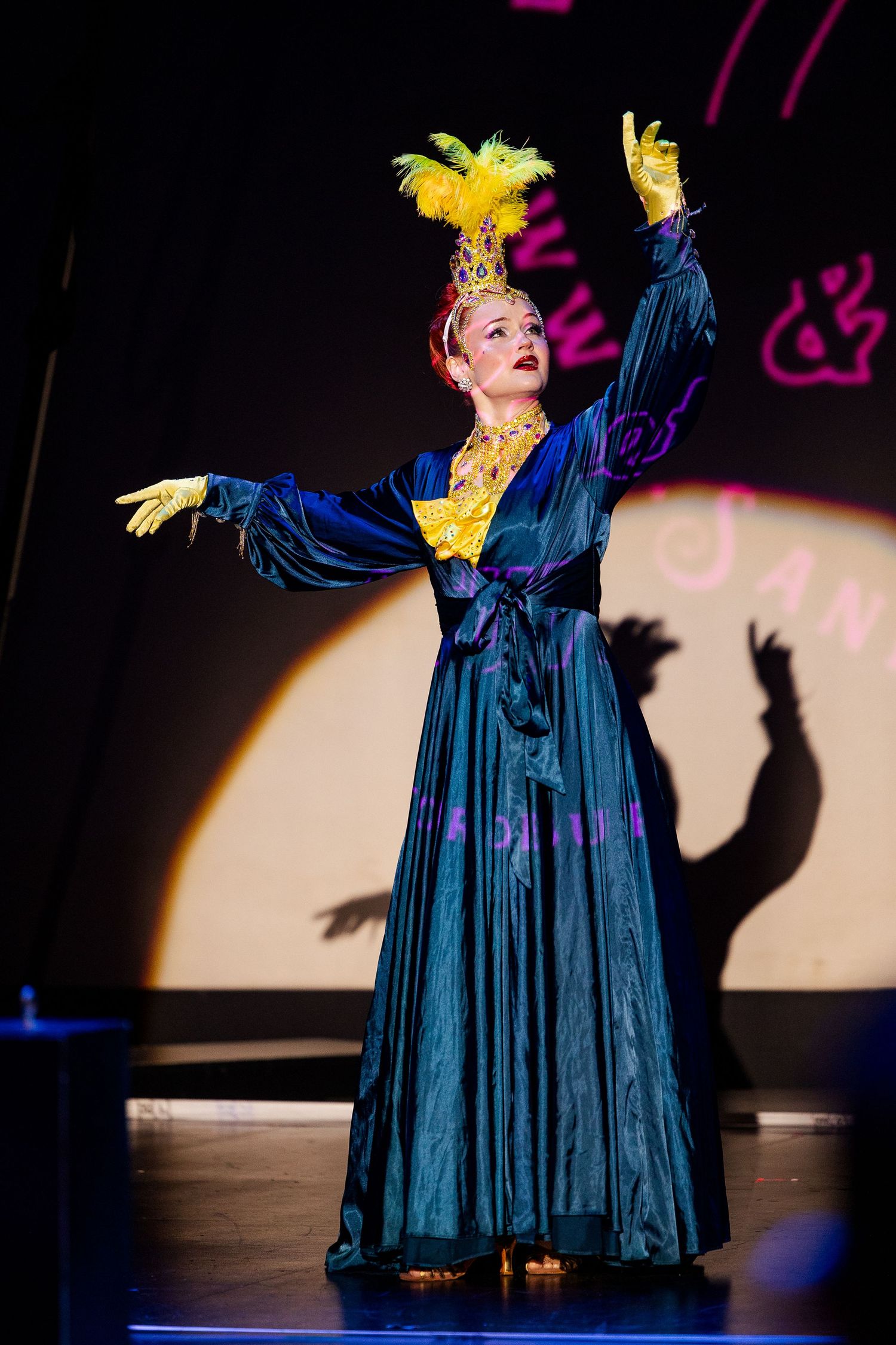 Burlesque dancer in teal robe and yellow feather crown poses with arms raised on illuminated stage.