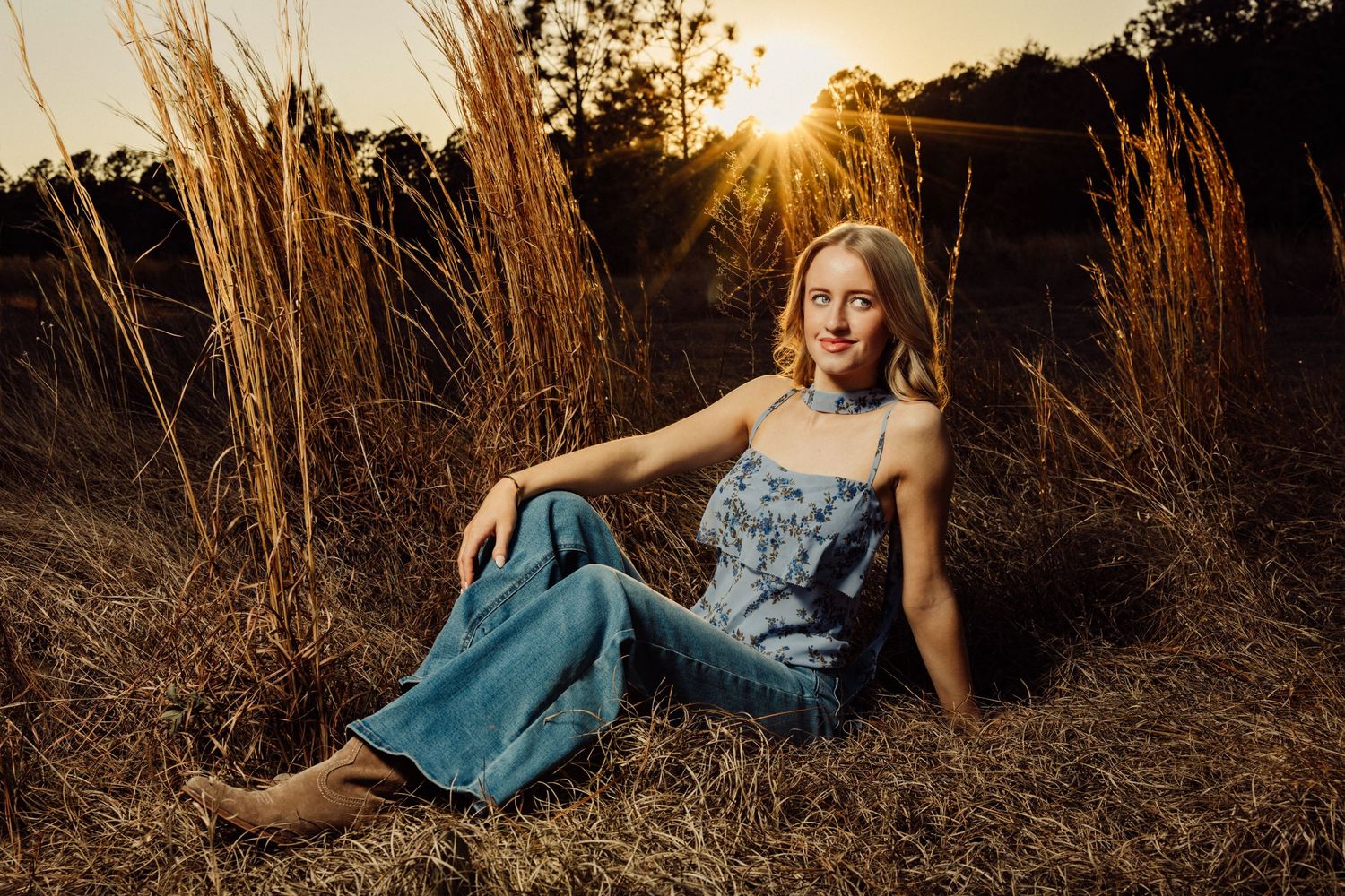 Young woman in denim overalls sitting in tall golden grass with sun flare at sunset.