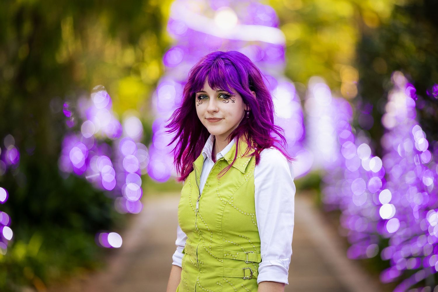 Young woman with vibrant purple hair wearing a green vest, posing in a bokeh-lit purple garden path.