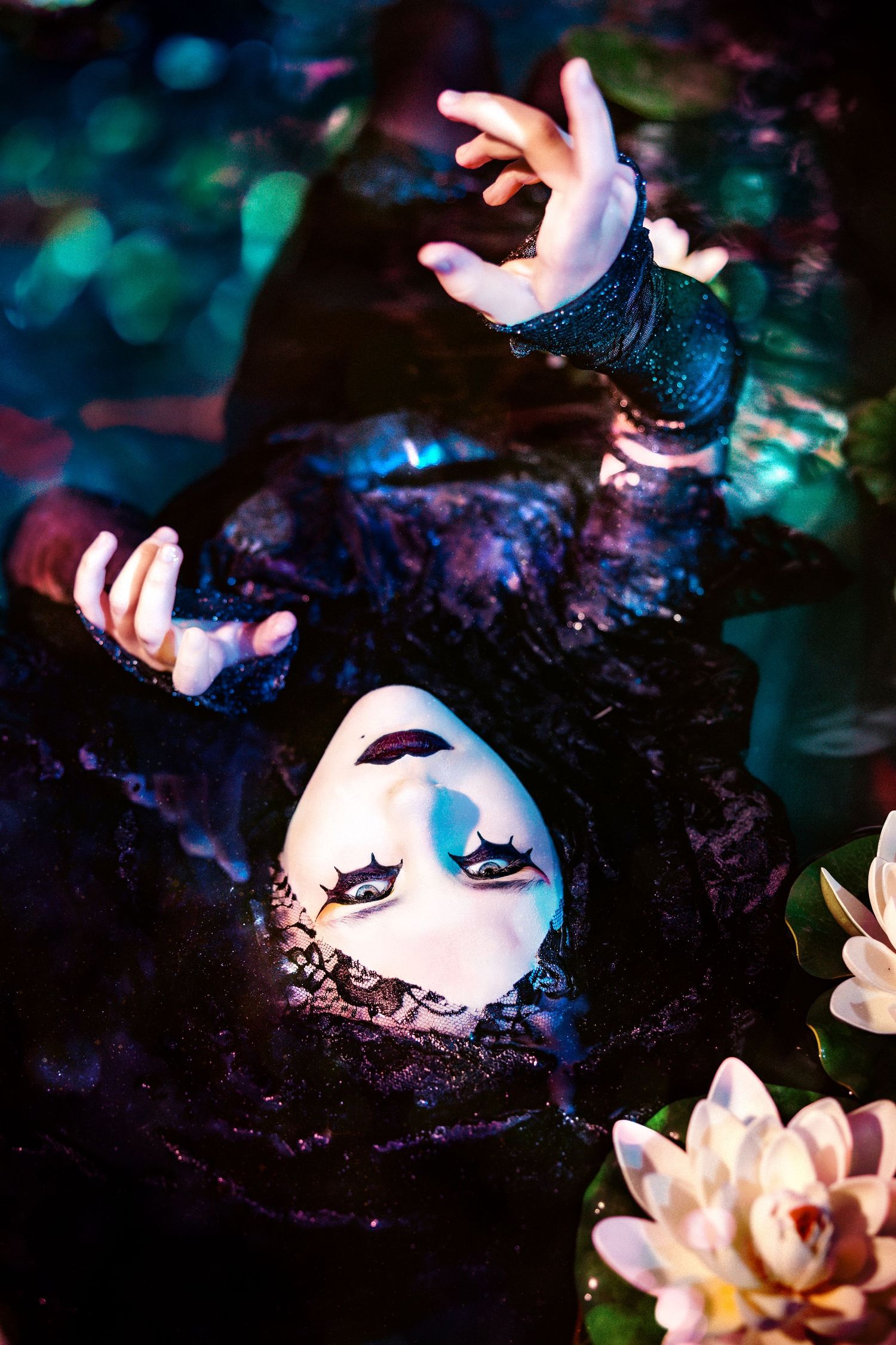 Woman in black lace with white mask reaching upward while floating in mystical lily pond.