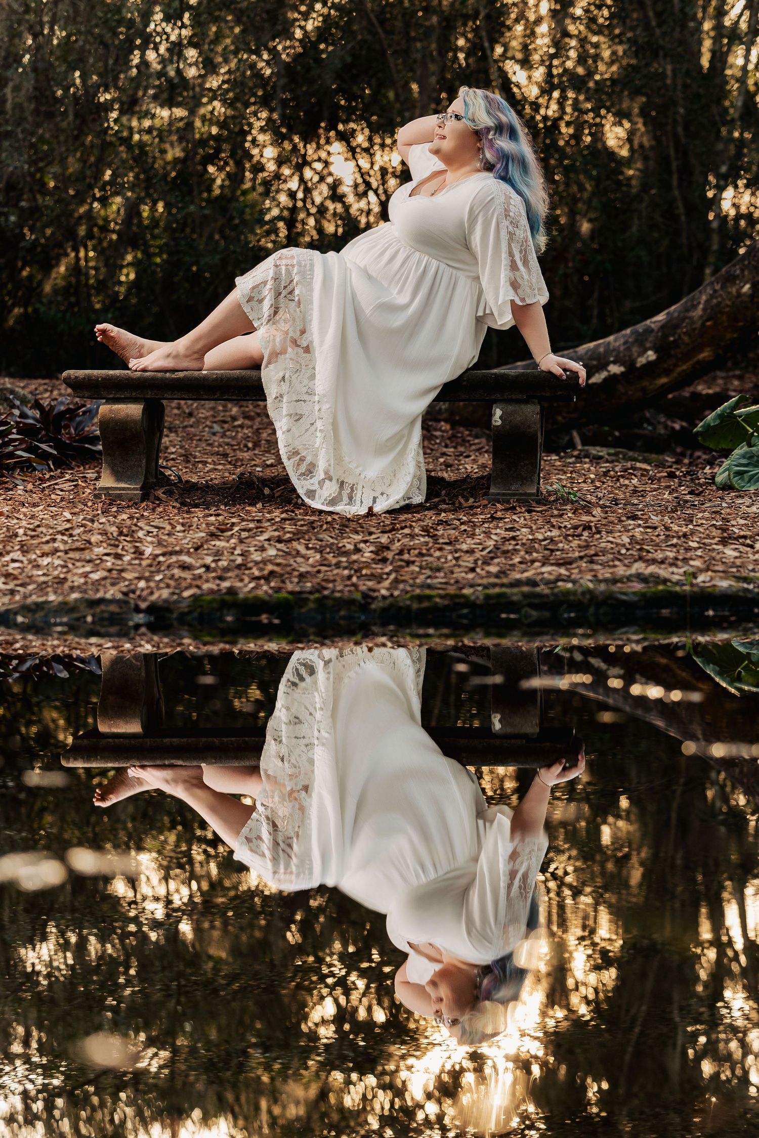Woman in white lace dress with blue hair reclines on bench, reflected in still water in forest setting.