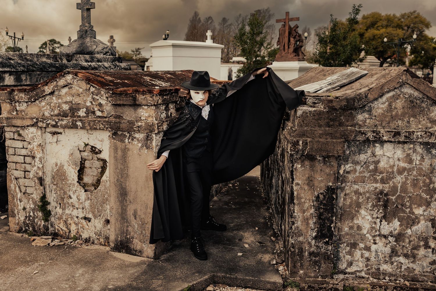 Dramatic caped figure in Venetian mask posing on weathered cemetery wall under stormy sky.