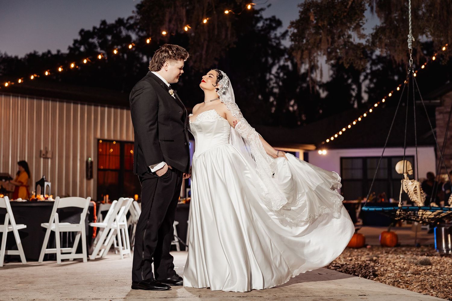Bride and groom share first dance outdoors at night under string lights, bride's gown flowing in the air.