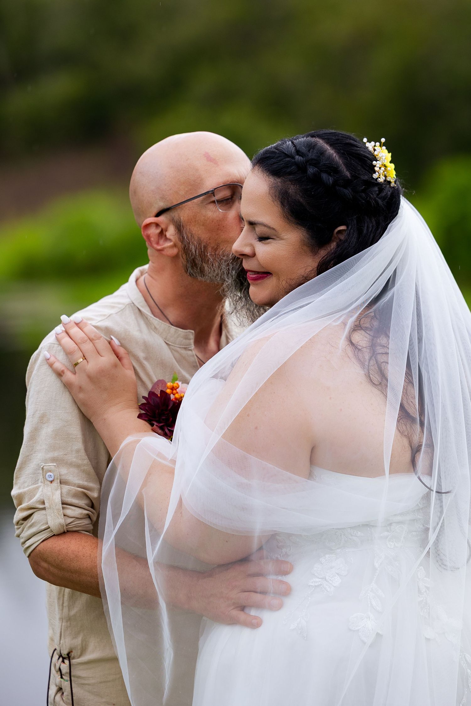 Bald groom tenderly kisses bride on cheek outdoors, bride wearing white veil and floral hair accessory.