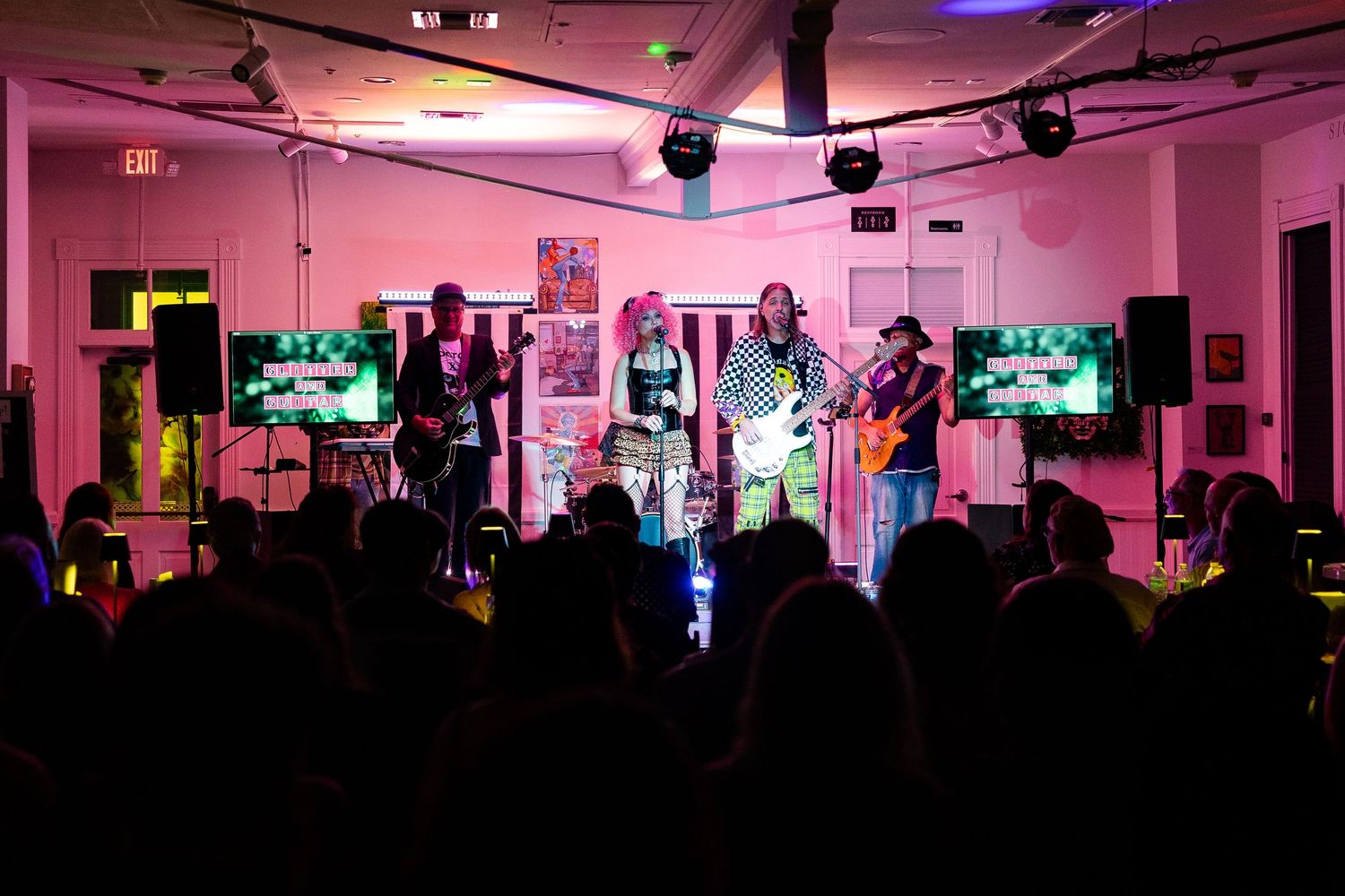Live band performing on stage with colorful pink lighting and audience silhouettes in a vibrant indoor venue.