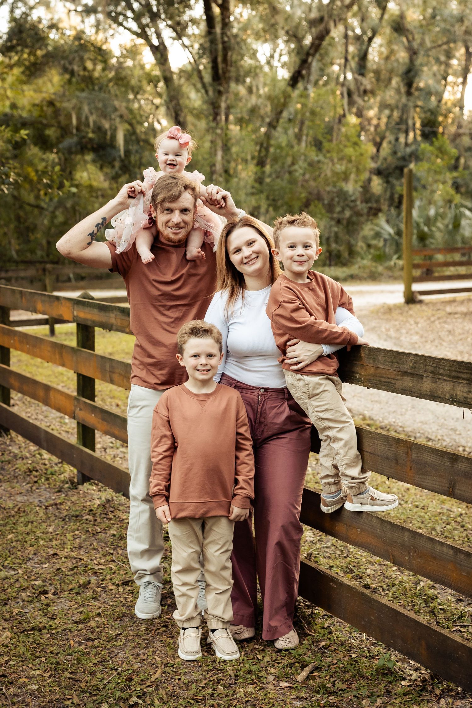 Happy family of five poses by a wooden fence in a lush green outdoor autumn setting.