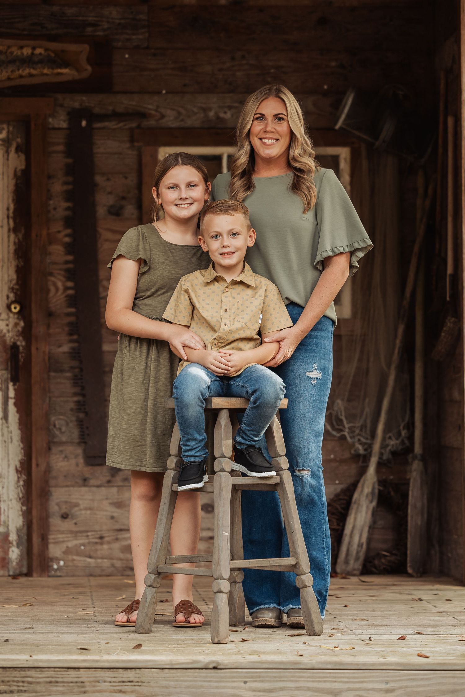 A smiling mother poses with her two children on a wooden stool in front of a rustic barn setting.