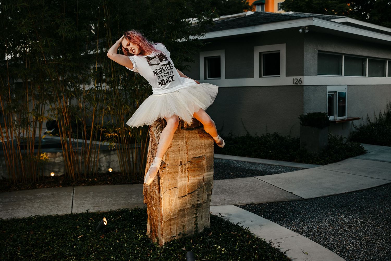 Ballerina in white tutu and graphic tee posing on a stone pedestal in a garden at dusk.