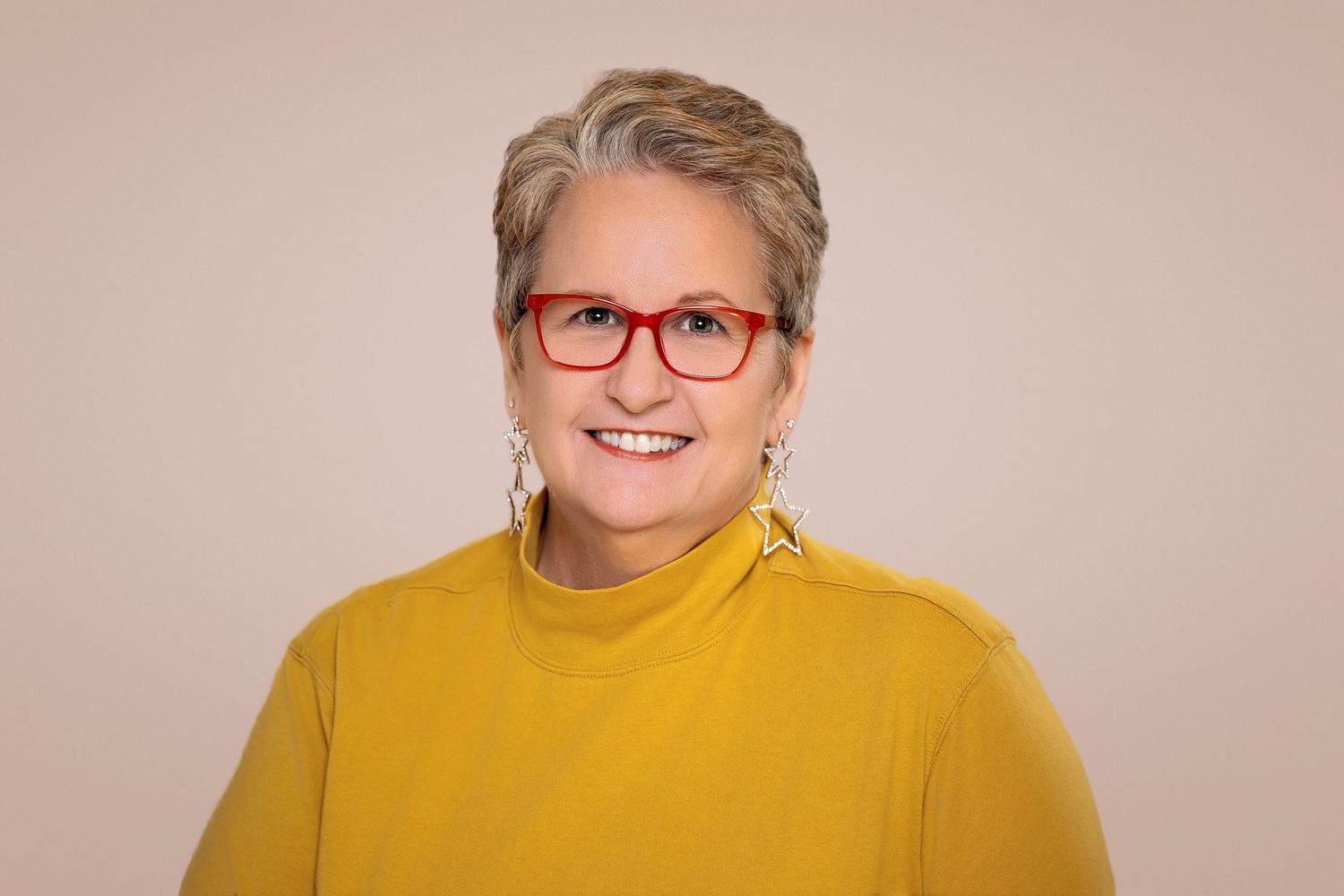 Smiling woman with short gray hair, red glasses, and yellow turtleneck sweater poses for a professional headshot.