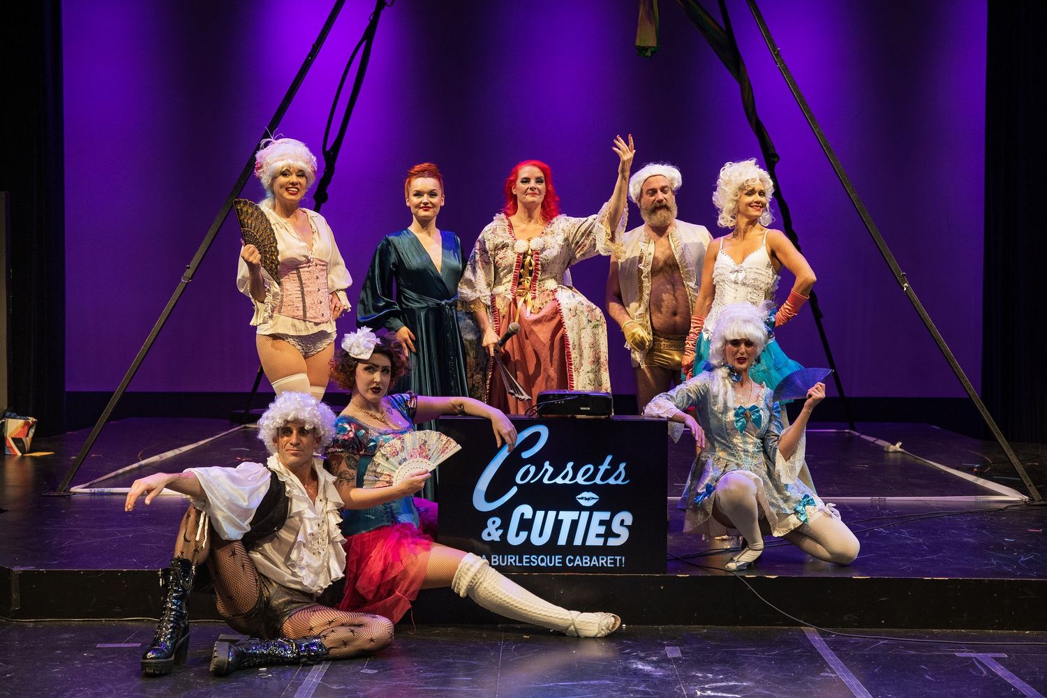 Corsets and Cuties burlesque cabaret performers pose together on stage in elaborate costumes.