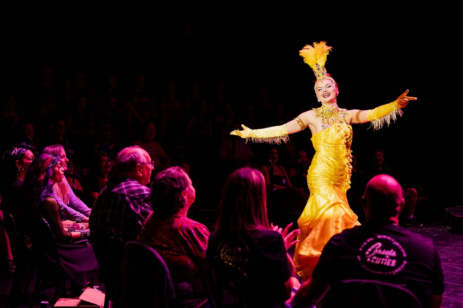 Burlesque performer in golden feathered gown dances before seated audience in dramatic stage lighting.