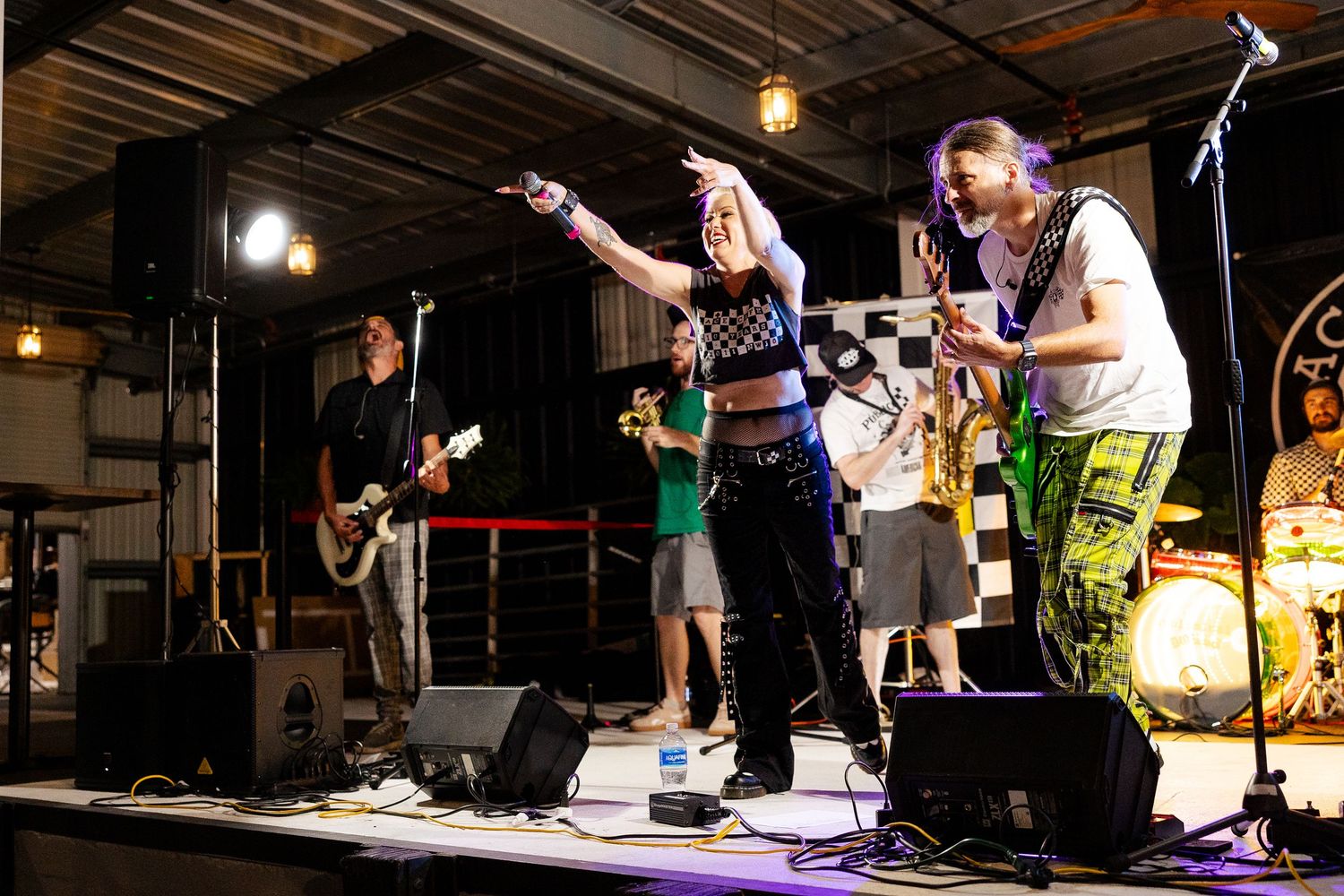 Band performing on stage with female vocalist pointing outward, guitarist, and drummer in industrial venue.