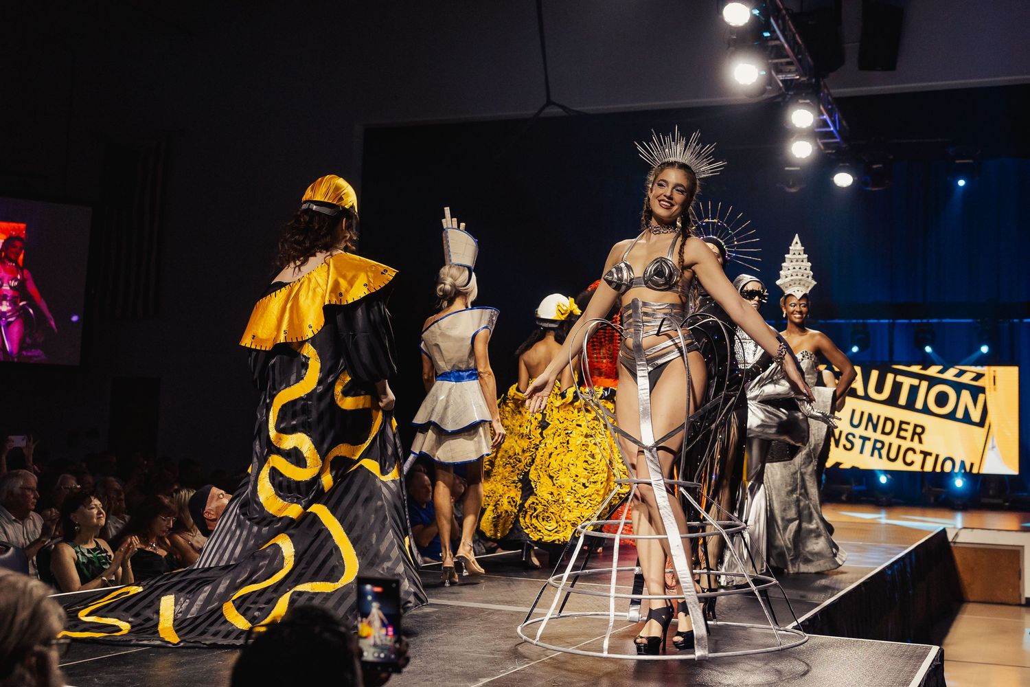 Models showcase avant-garde construction-themed fashion on runway with Caution Under Construction sign.