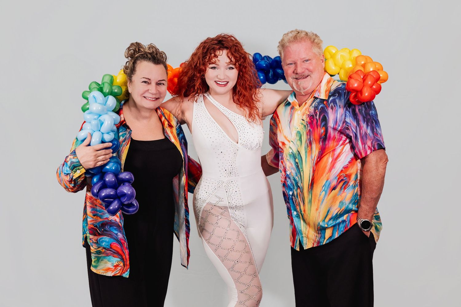 Three people pose together, two wearing rainbow balloon shoulder pieces and one in a white lace outfit.