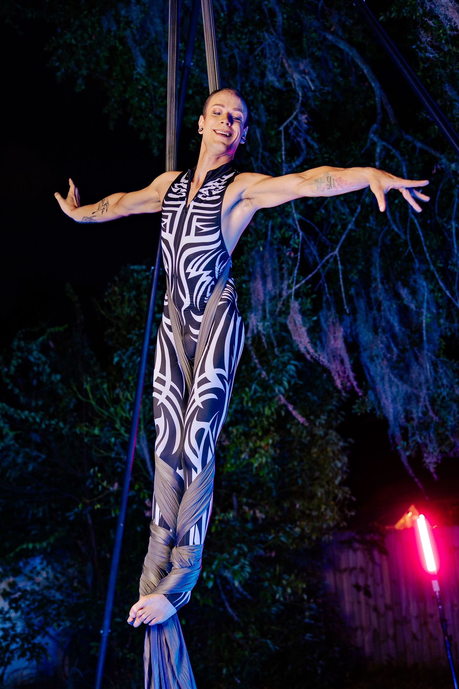 Aerial silk performer in black and white tribal costume balances gracefully with arms outstretched outdoors at night.