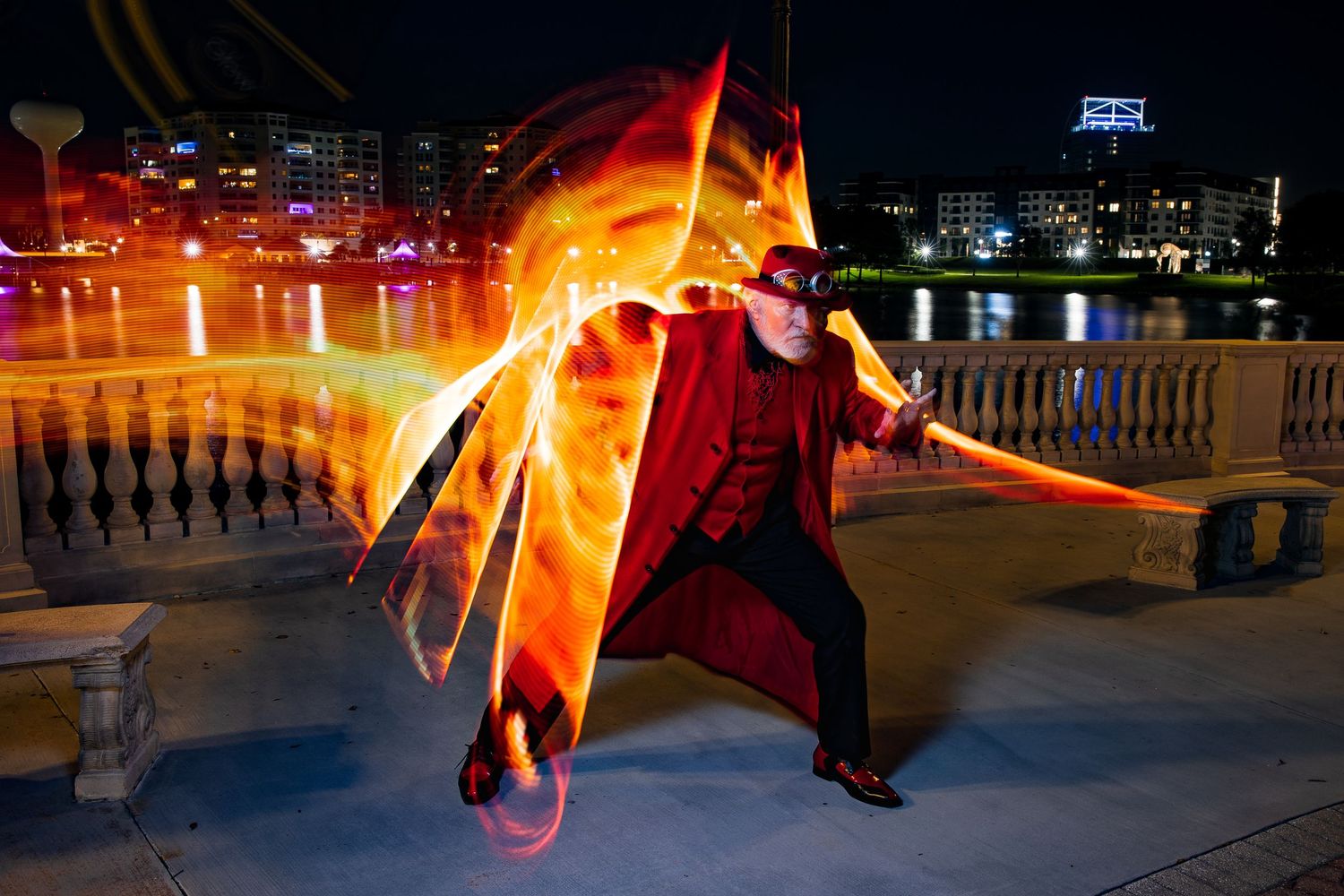 Man in red costume leaps dynamically with glowing orange light trails at night by waterfront.