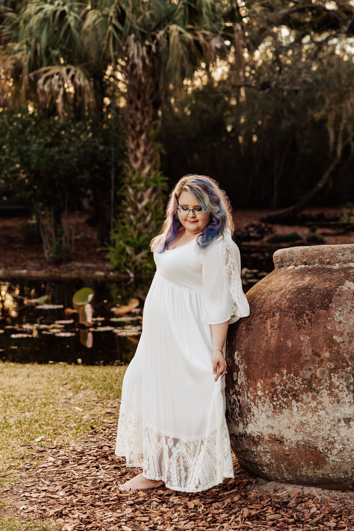 Woman in white lace maxi dress with colorful hair and glasses poses beside rustic urn in lush garden setting.